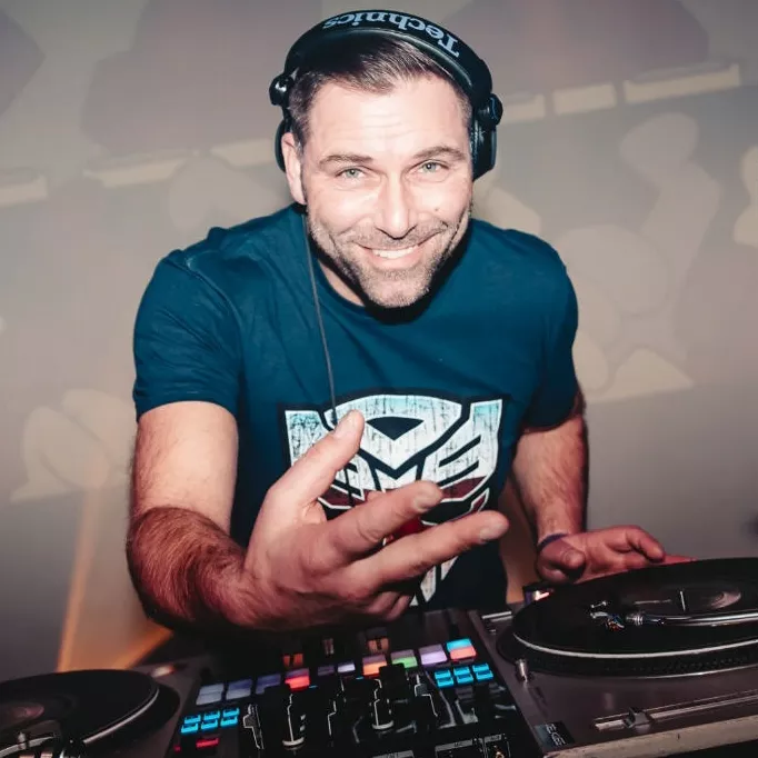 DJ DNS (Dennis Bruijn) draait glimlachend achter de draaitafels met koptelefoon op en een shirt met het Transformers-logo, terwijl hij het publiek aanspreekt in een energieke clubsfeer.